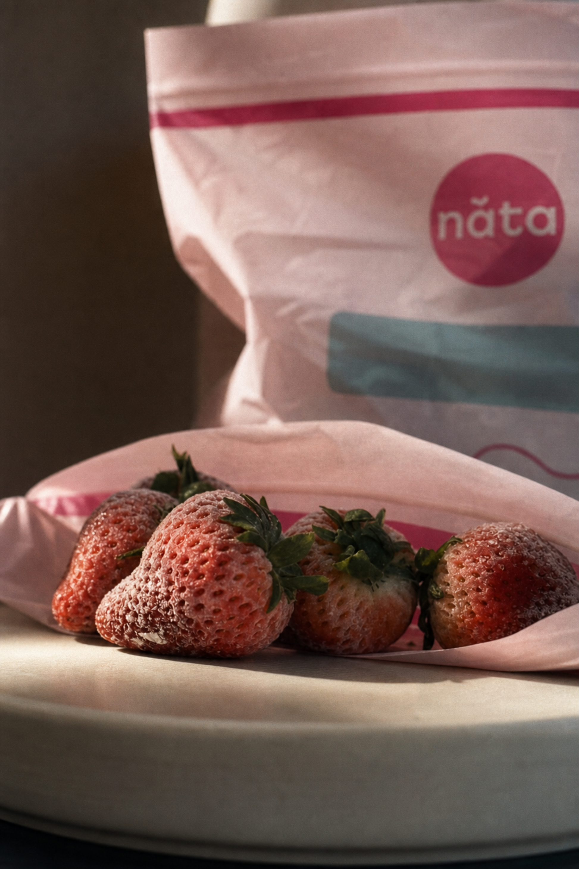 Fresh strawberries resting on a pink paper liner in the foreground, with a branded Năta bag blurred in the background on a dark surface.