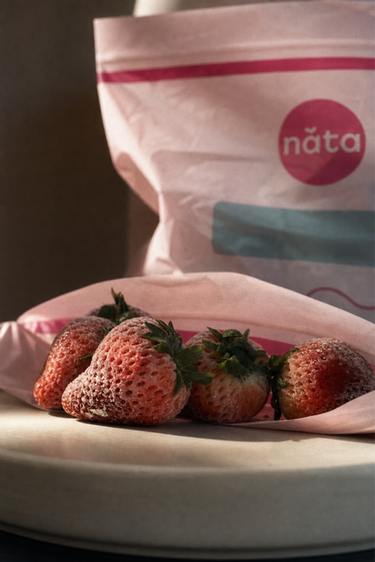 Fresh strawberries resting on a pink paper liner in the foreground, with a branded Năta bag blurred in the background on a dark surface.